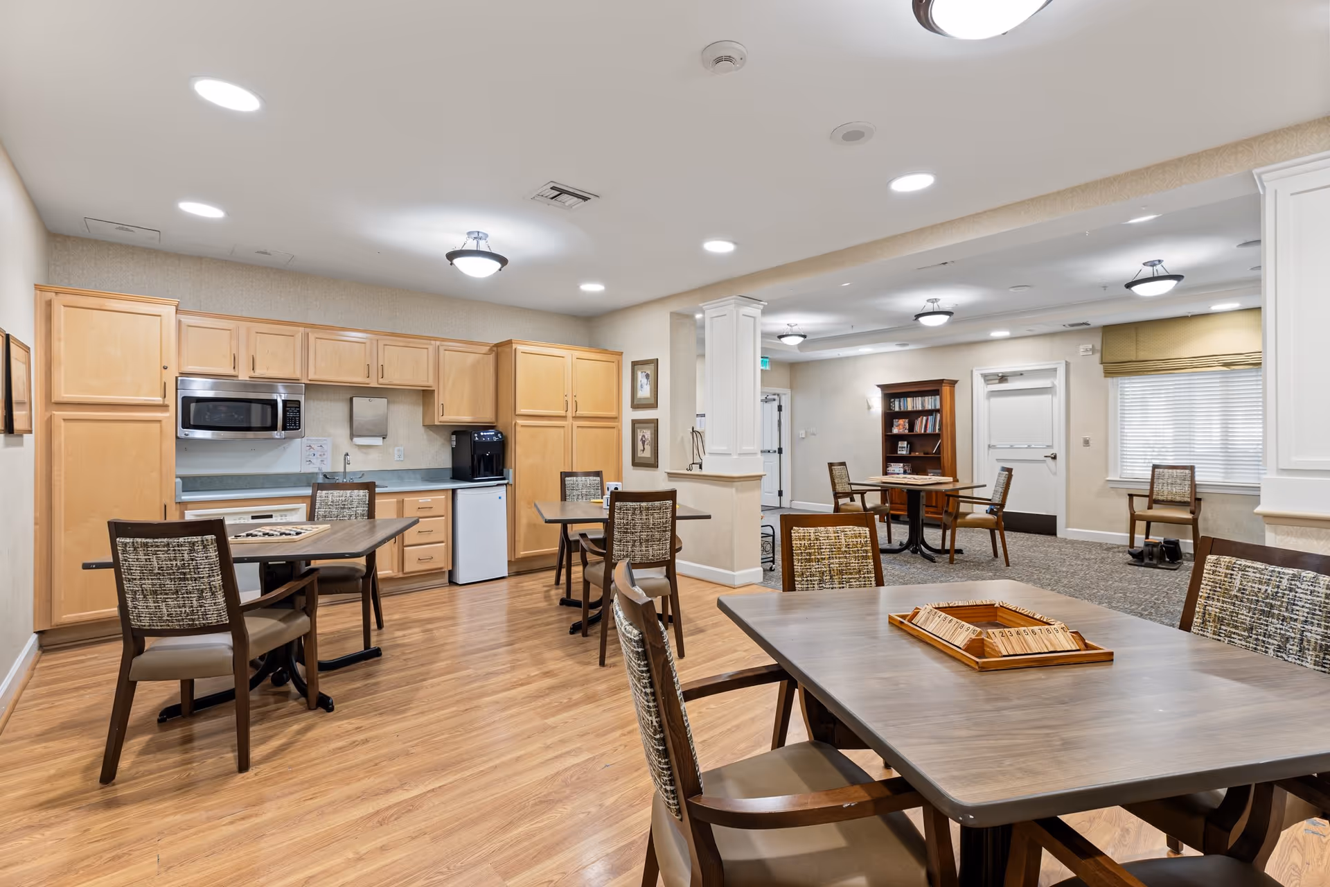 Well-lit communal dining and kitchenette area with multiple tables and chairs, light wood cabinets, and seating.