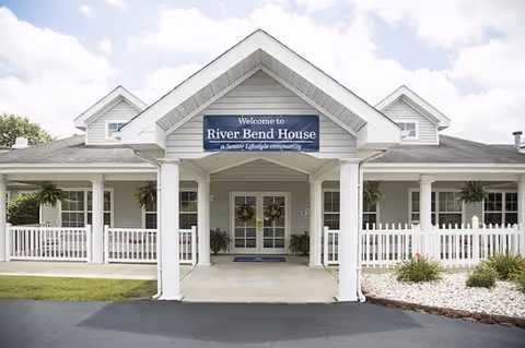 Front exterior view of a single-story senior living community building with a covered entrance supported by white columns. A sign above the entrance reads 'Welcome to River Bend House, a Senior Lifestyle community.' The building has light gray siding, white trim, and a white picket fence along the front porch. There are potted plants and hanging ferns decorating the porch area.