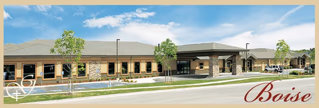 Exterior view of a single-story assisted living facility building with a covered entrance, stone and beige siding, several windows, small trees, and a parking area in front under a blue sky with some clouds.