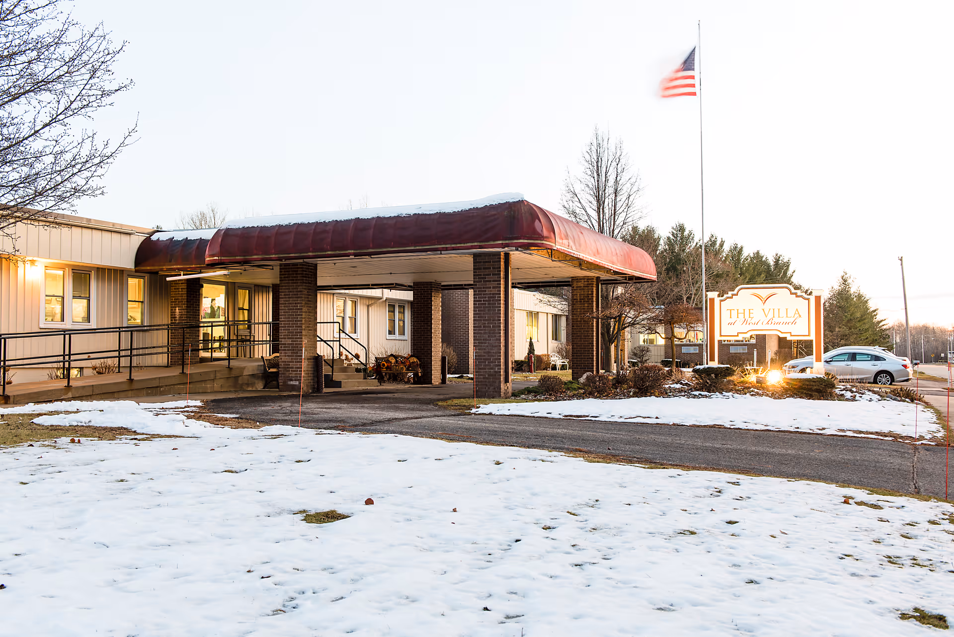 Exterior view of The Villa at West Branch senior living facility during winter with snow on the ground, a covered entrance with brick pillars, an American flag on a flagpole, and a sign displaying the facility name.