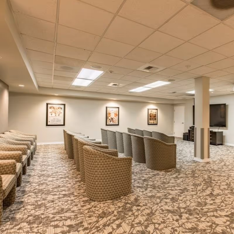 A large room with multiple rows of cushioned chairs arranged facing a wall-mounted flat screen TV. The room has a patterned carpet, a drop ceiling with fluorescent lighting, and framed pictures on the walls.