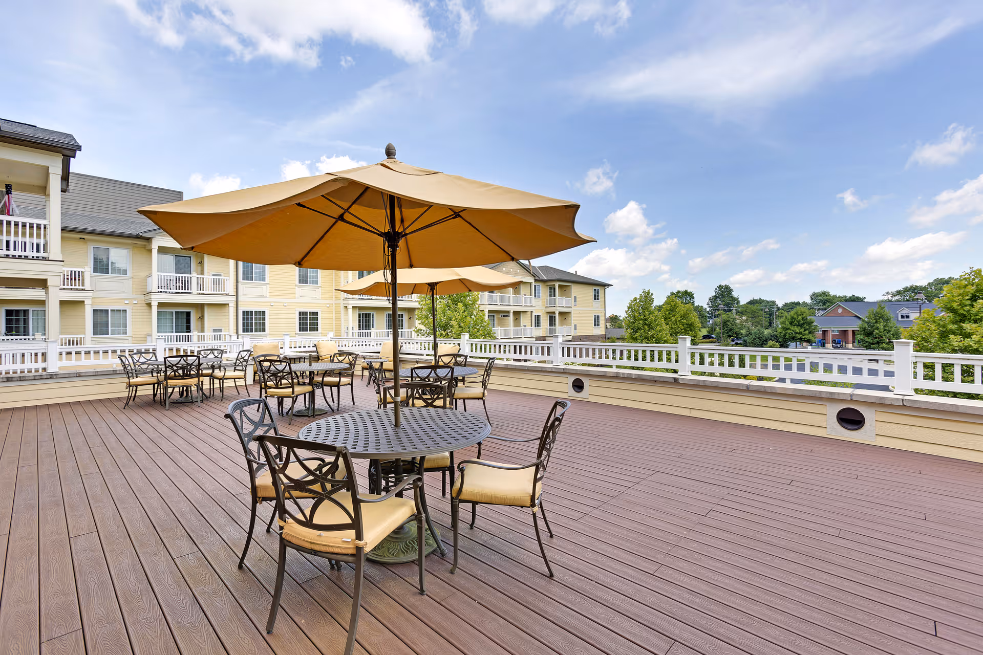 Outdoor patio area with multiple round metal tables and chairs with beige cushions, shaded by large beige umbrellas. The patio has wooden flooring and white railing, with a multi-story residential building and trees in the background under a partly cloudy sky.