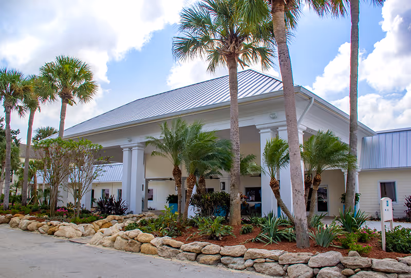 Exterior view of Buena Vida Estates senior living facility with a covered entrance, white columns, palm trees, and landscaped garden beds with rocks and various plants under a partly cloudy sky.