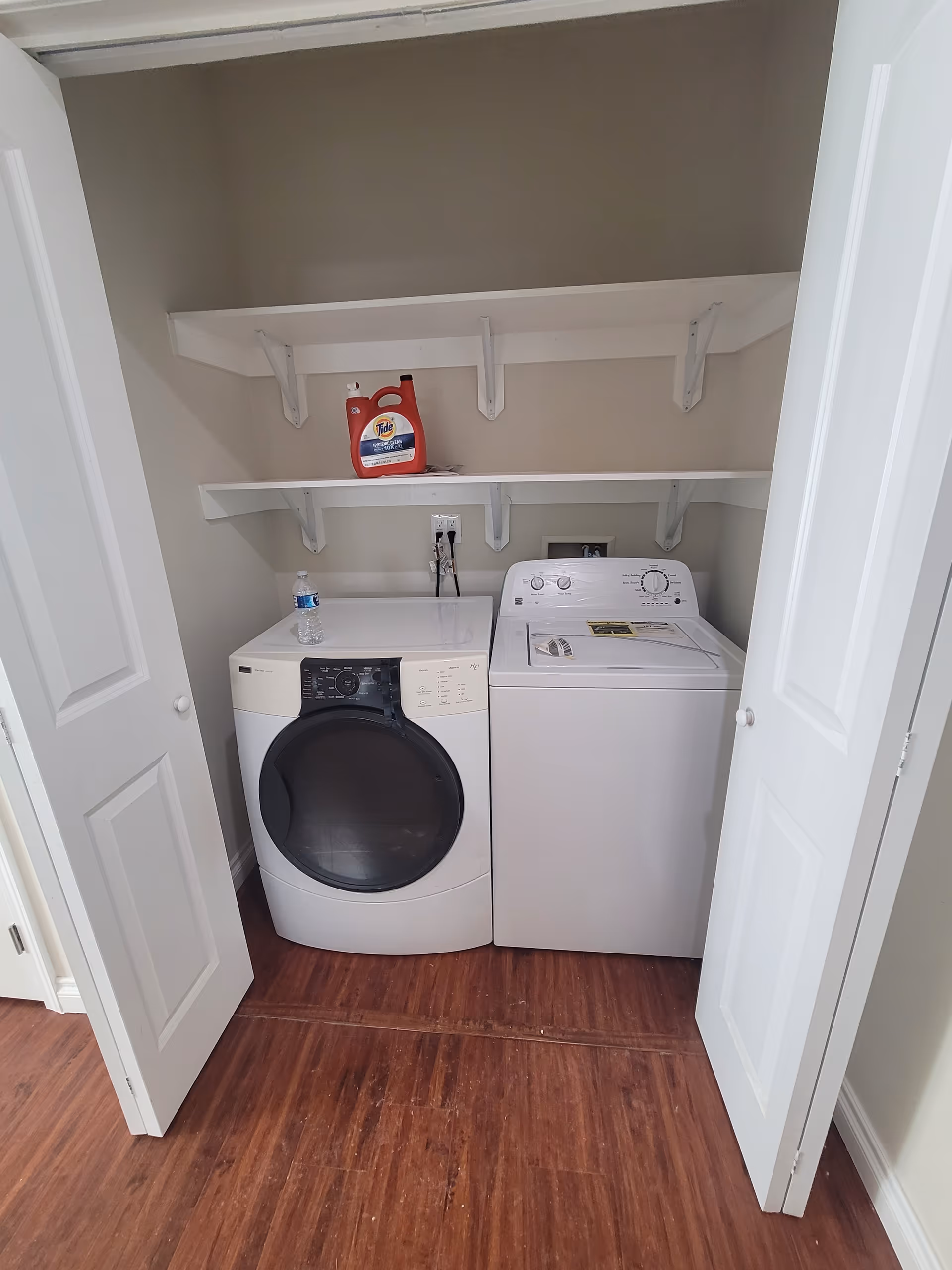 Laundry area with a front-loading washing machine and a top-loading dryer side by side inside a closet with white double doors. Two white shelves above hold a bottle of Tide detergent and a water bottle. The floor is wooden and the walls are painted beige.