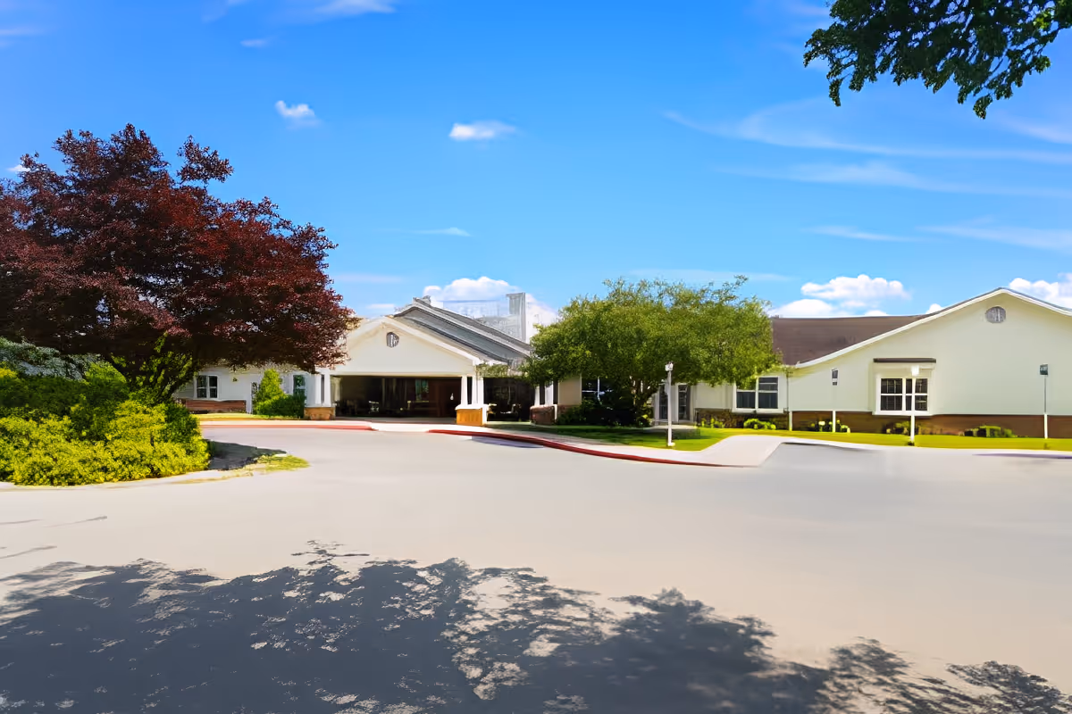 Front exterior view of a single-story senior living facility building with a covered entrance, surrounded by trees and greenery under a bright blue sky with a few clouds.