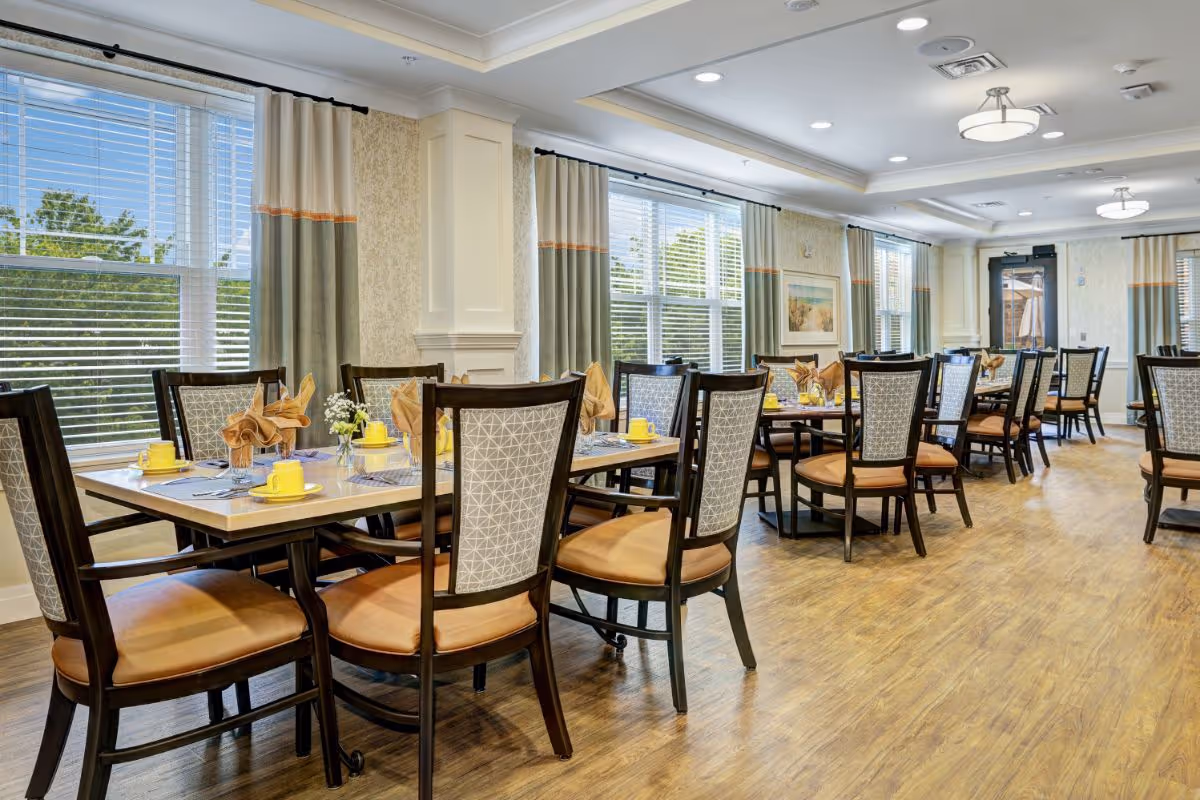 Bright communal dining room with multiple set tables and chairs, large windows with curtains, and wood flooring.