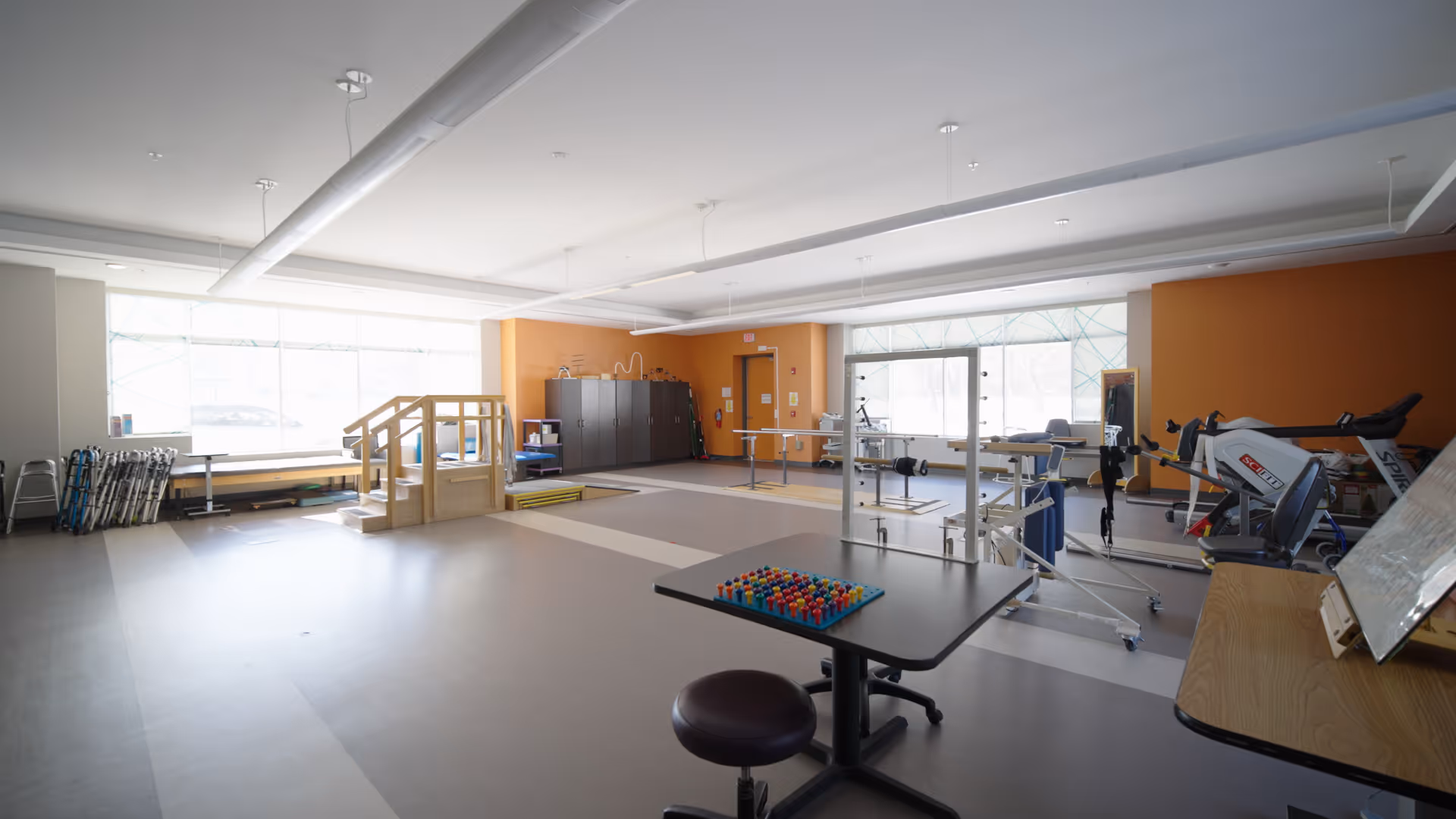 A spacious rehabilitation room with exercise equipment, parallel bars, a small staircase for physical therapy, and a table with a pegboard game. The room has large windows allowing natural light and orange accent walls.