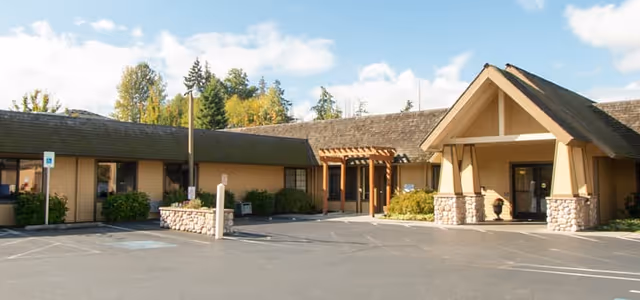 Single-story senior living facility building with a covered entrance, parking lot, and landscaping under a partly cloudy sky.