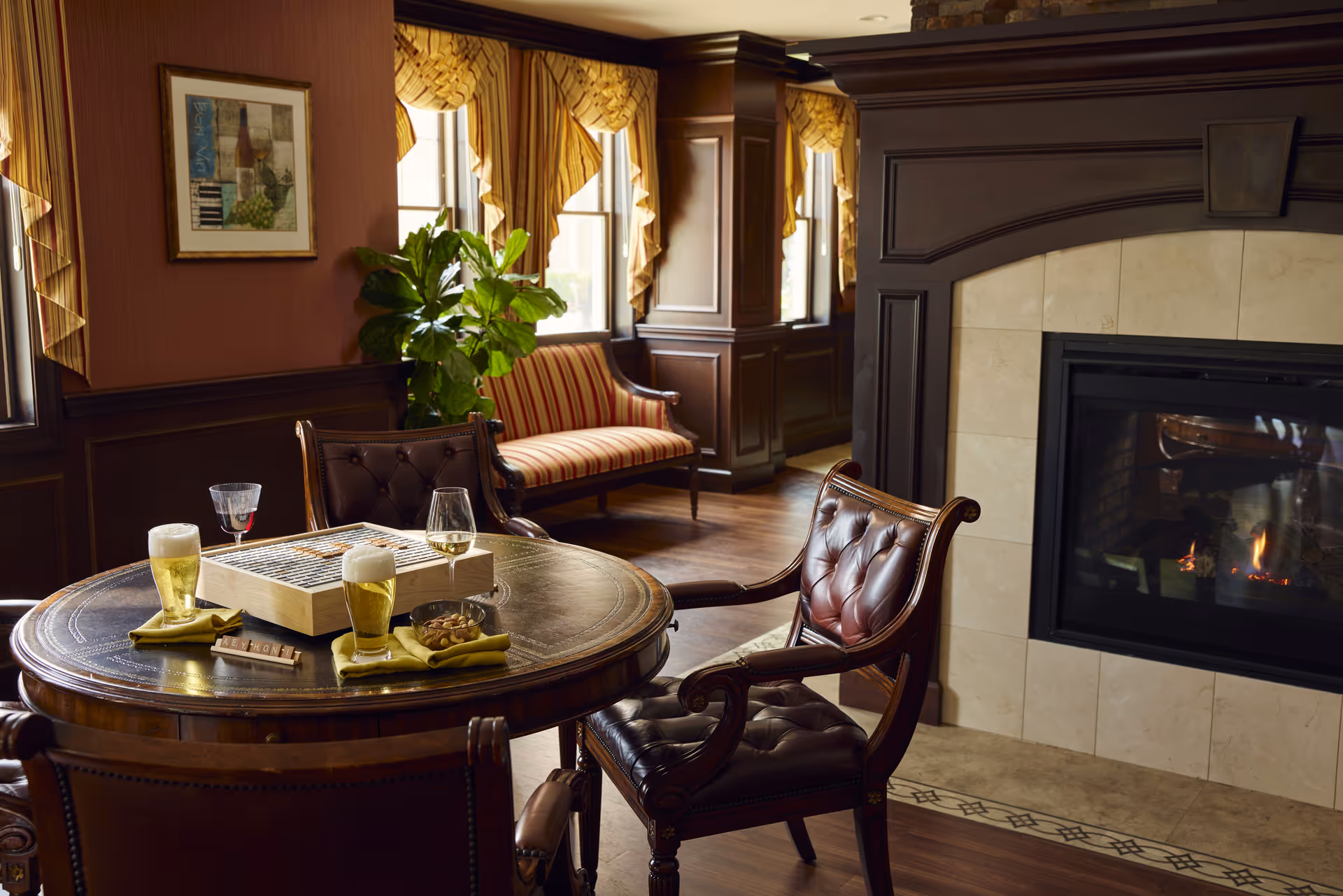 Cozy lounge with a round table holding drinks and a board game, tufted leather chairs, a striped settee by draped windows, and a lit fireplace.