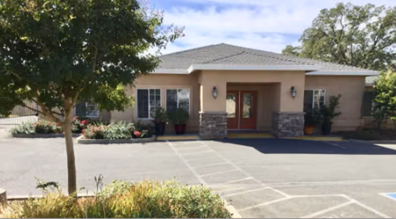 Front exterior view of a single-story building with a covered entrance supported by stone pillars, surrounded by landscaped plants and trees, with a parking area in front.