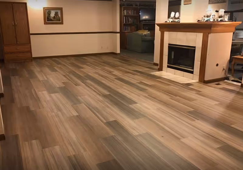 An empty room with wood-patterned flooring, a fireplace with a wooden mantle, a built-in wooden cabinet, and a small framed picture on the wall. In the background, there is a glimpse of a living area with a couch and a television.
