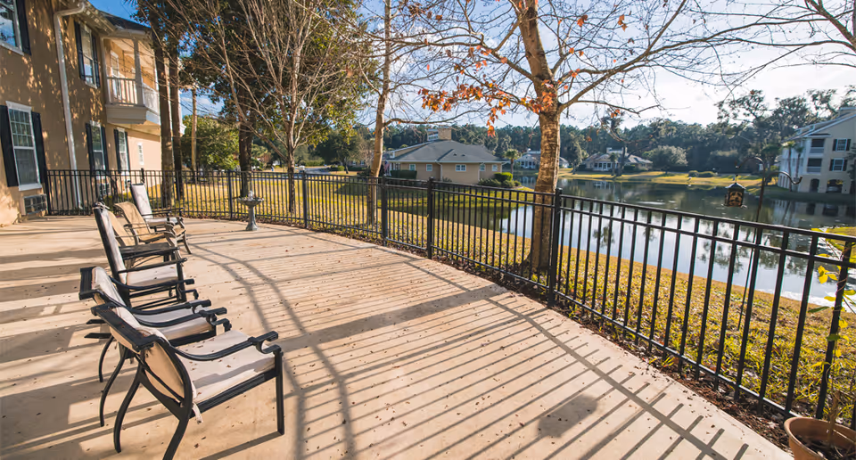 Outdoor patio area with several empty chairs lined up facing a black metal railing overlooking a pond and trees, with residential buildings in the background under a clear sky.