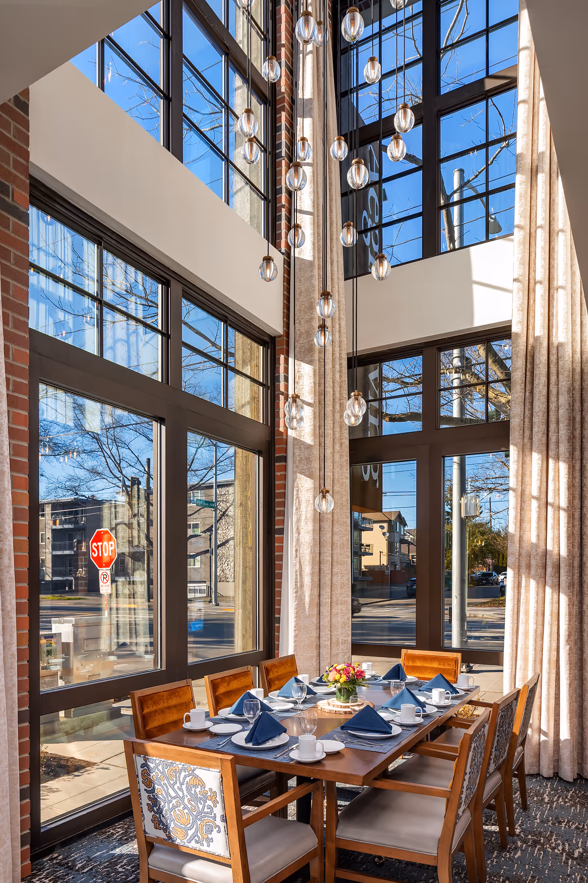 A bright dining room with a wooden table set for eight people, featuring blue napkins, white plates, cups, and a centerpiece of colorful flowers. The room has large floor-to-ceiling windows with beige curtains, allowing natural light to flood in. A modern chandelier with multiple hanging glass bulbs is suspended from the high ceiling.