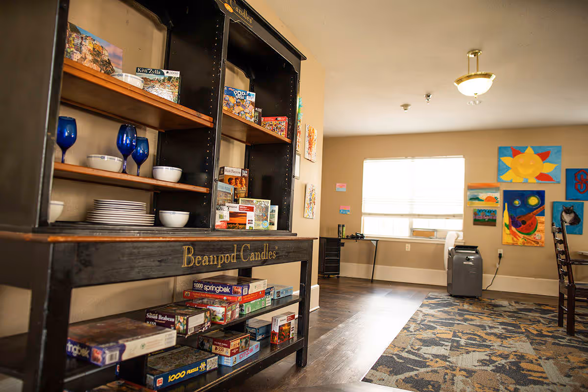 Interior view of a room with a black wooden shelf labeled 'Beanpod Candles' holding blue wine glasses, white bowls, plates, and several puzzle boxes. The room has beige walls, a window with blinds, colorful artwork on the walls, a patterned rug on the floor, and a small desk with a chair near the window.