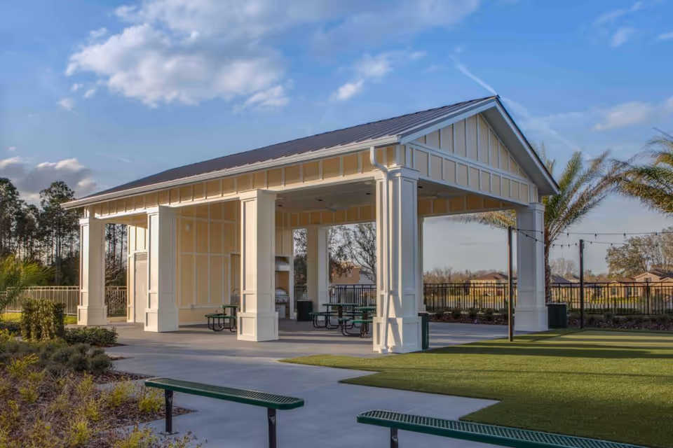 Outdoor covered pavilion with picnic tables and benches on a concrete patio surrounded by landscaped greenery and palm trees under a partly cloudy sky.