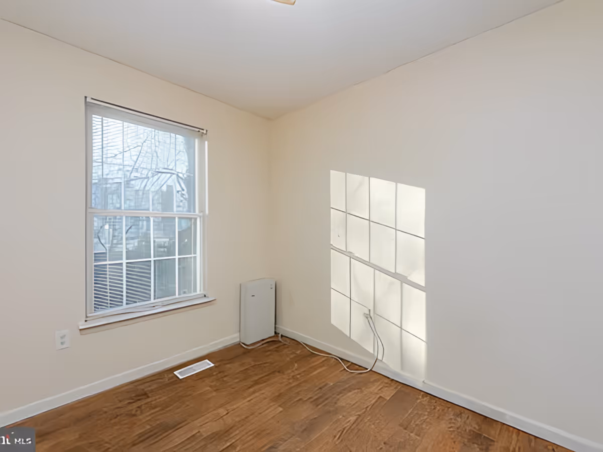 Empty room with beige walls, a window with blinds partially open, wooden floor, and a small white heater or air purifier in the corner. Sunlight casts a grid pattern shadow on the adjacent wall.
