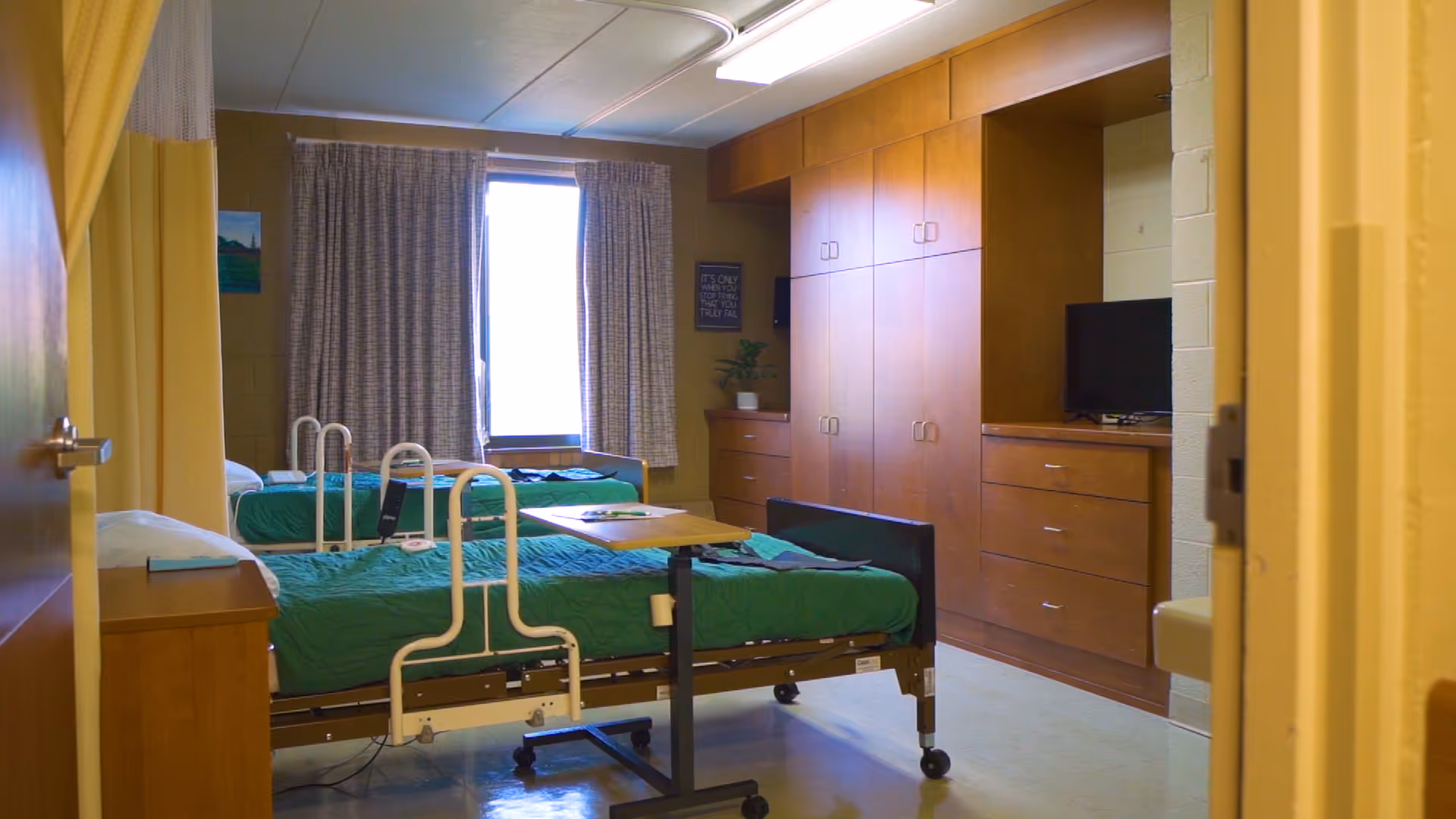 A room with two hospital-style beds with green bedding, a small overbed table, wooden cabinets and drawers along one wall, a window with curtains, and a television on a cabinet.