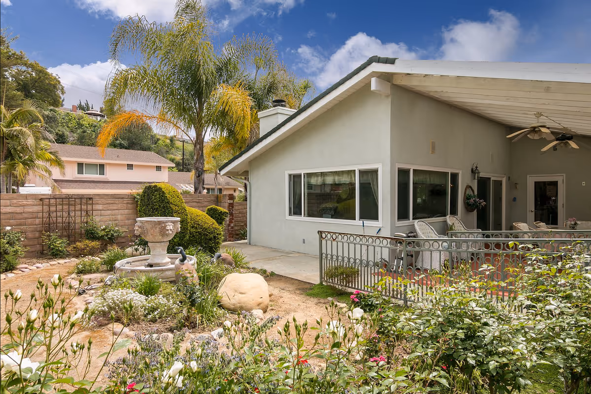 Outdoor garden area with various plants and flowers, a stone birdbath fountain, and trimmed bushes. In the background, there is a single-story building with a covered patio featuring ceiling fans and outdoor seating. The sky is partly cloudy with blue patches.
