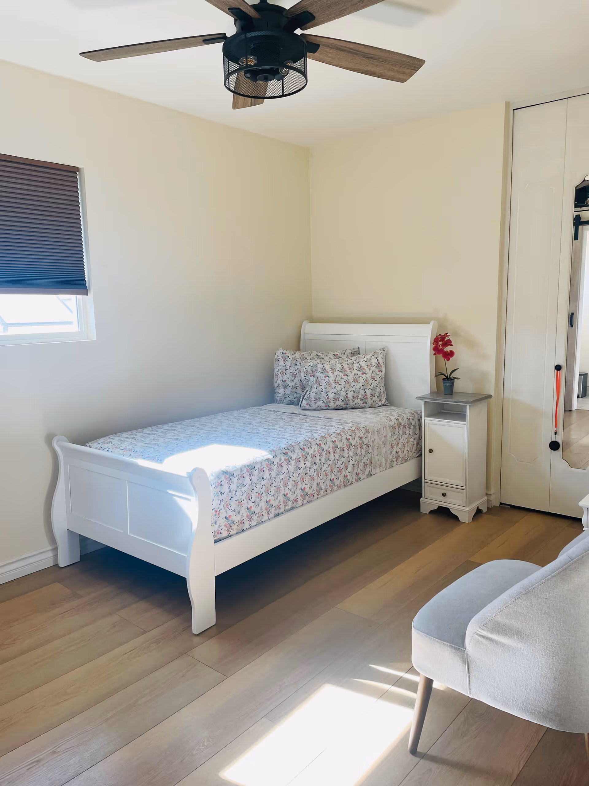 A bright white bedroom with a single bed, floral bedding, a white nightstand with a small plant, window with blinds, a ceiling fan, and light wood flooring.