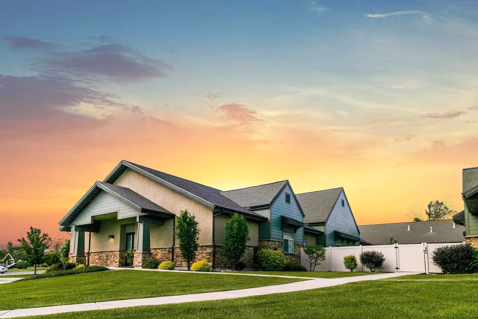 Single-story senior living building with pitched roofs, landscaped lawn, and a colorful sunset sky.