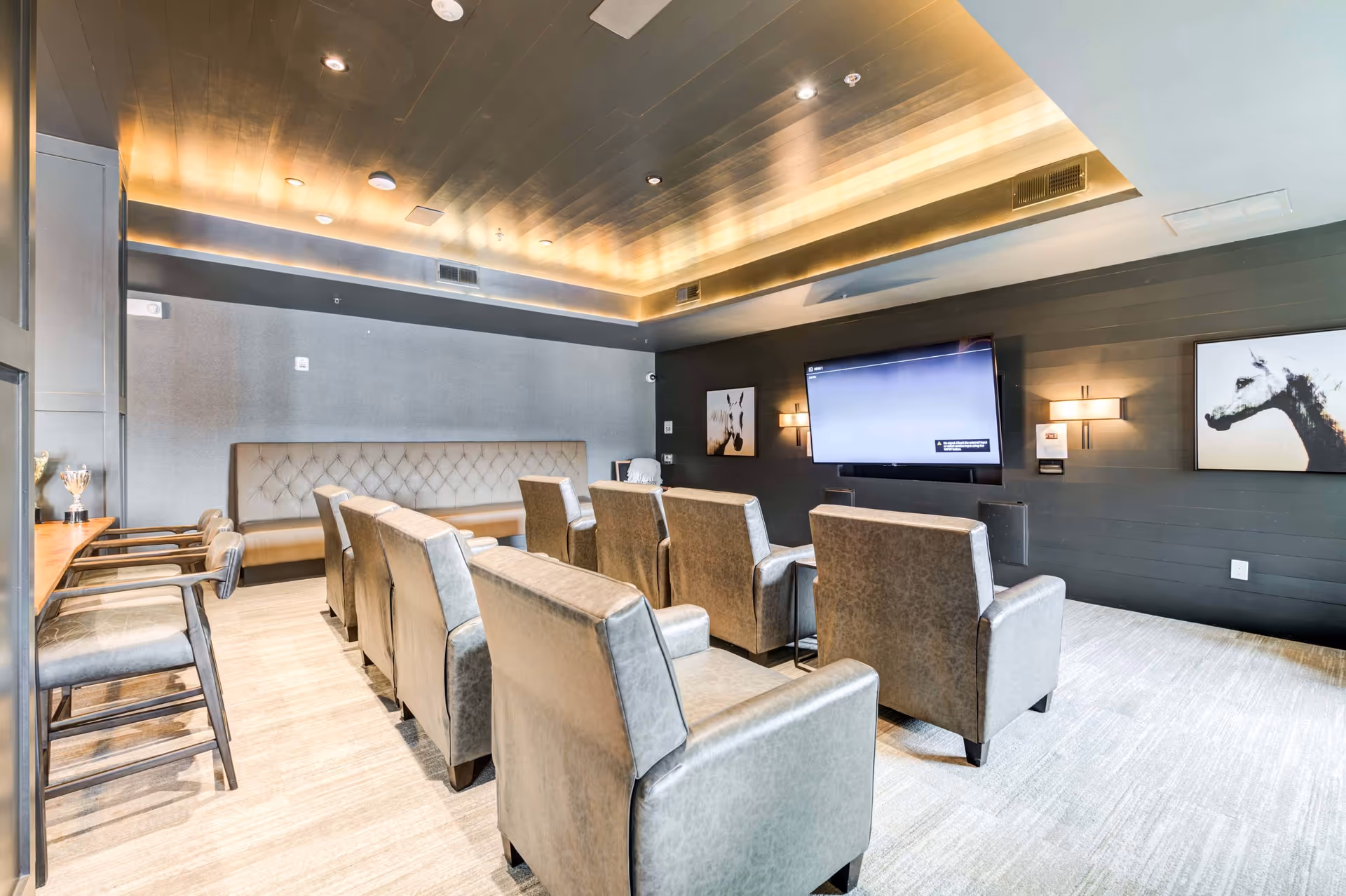 A modern media room with several rows of gray upholstered chairs facing a large flat-screen TV mounted on a dark wood-paneled wall. The room features a tufted bench along the back wall, two framed animal artworks on either side of the TV, and a ceiling with recessed lighting and a warm glow. There are also a few bar stools and a counter on the left side of the room.