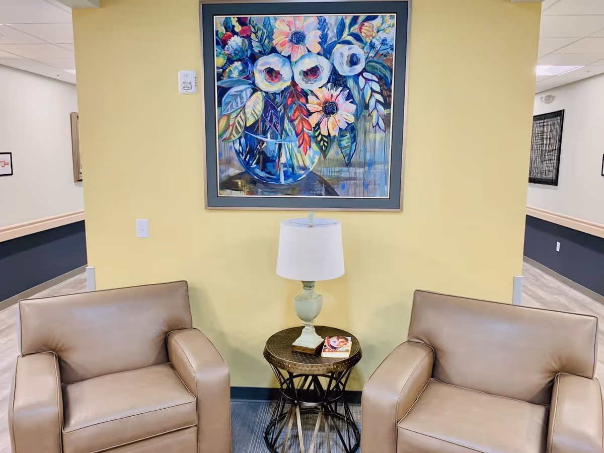 Two tan leather armchairs with a small round metal table between them, holding a white table lamp and a book, set against a yellow wall with a colorful floral painting hanging above.
