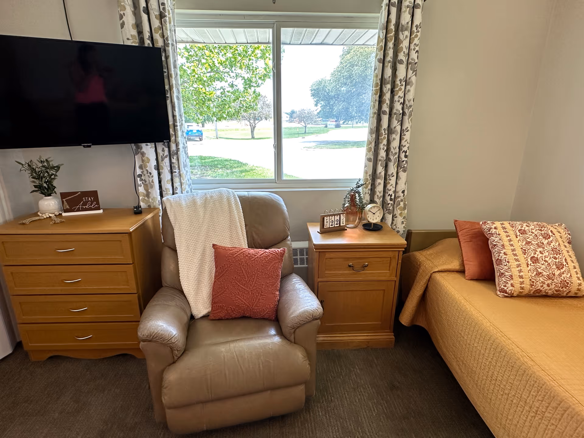 Cozy senior living bedroom with a recliner, wooden dresser and nightstand next to a single bed under a window looking out to trees.
