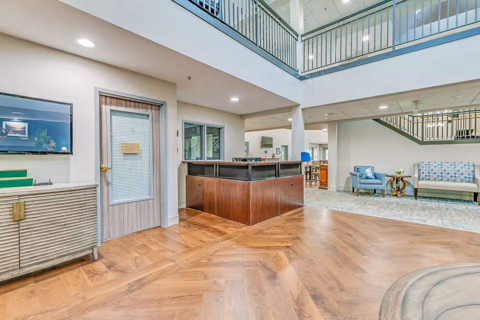 Interior view of a senior living facility lobby with a wooden reception desk in the center, a door with a frosted glass panel on the left, seating area with a blue armchair and a patterned bench on the right, and a staircase with metal railings leading to the upper floor.