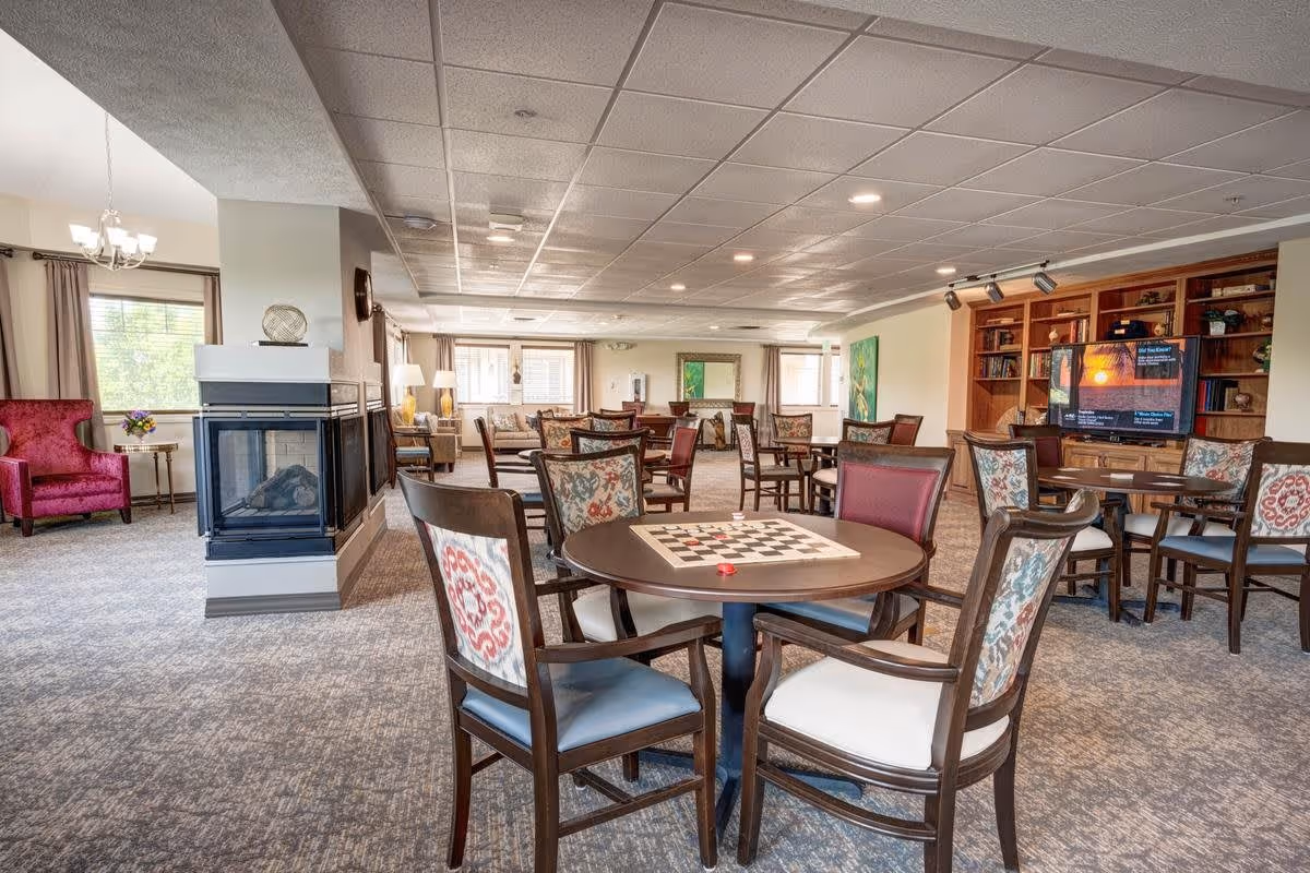 A spacious senior living common area with multiple round tables and chairs, some with checkerboards on them. There is a fireplace on the left side, a red armchair near a window, and a large wooden bookshelf with a TV on the right. The room is well-lit with ceiling lights and natural light from windows.