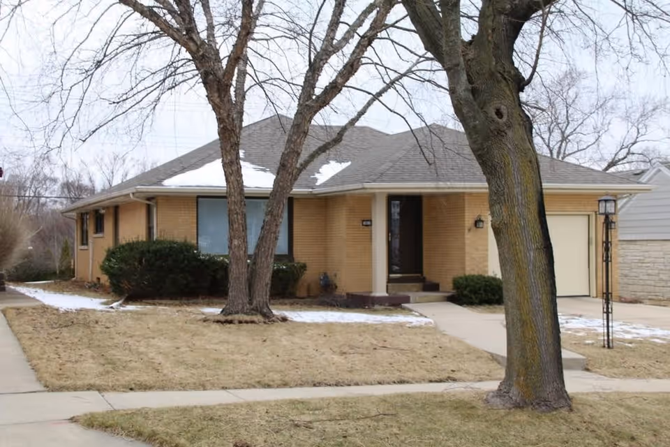 Single-story brick residential building with a sloped roof partially covered in snow, a front porch with steps, a garage, and leafless trees in the front yard during winter.
