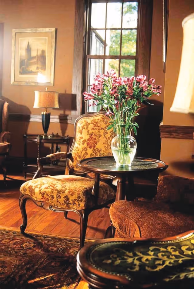A cozy living room area with vintage-style upholstered chairs and a small round wooden table holding a glass vase filled with pink flowers. The room features warm brown walls, a window letting in natural light, a framed picture on the wall, and a table lamp on a side table.