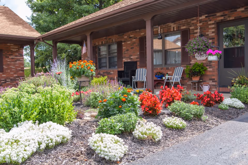 Brick-front porch with a covered walkway, two chairs and a colorful landscaped flower garden in front.