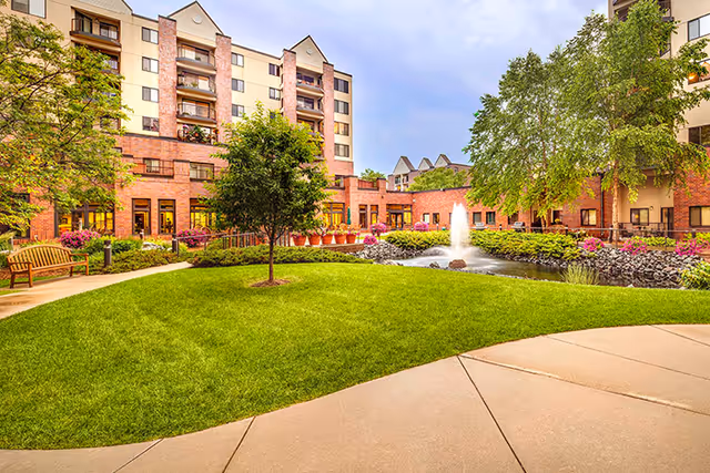 Outdoor courtyard area of a senior living facility featuring a well-maintained green lawn, a small tree, a water fountain in a pond surrounded by rocks and flowers, benches, and a multi-story building with balconies and large windows in the background under a partly cloudy sky.