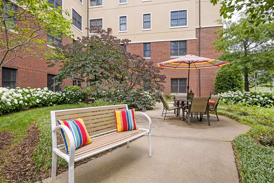 Outdoor patio area at Brookdale Green Hills Cumberland featuring a white bench with colorful striped cushions, a round table with chairs, and a large striped umbrella. The area is surrounded by green grass, bushes with white flowers, and trees, with a multi-story brick and beige building in the background.