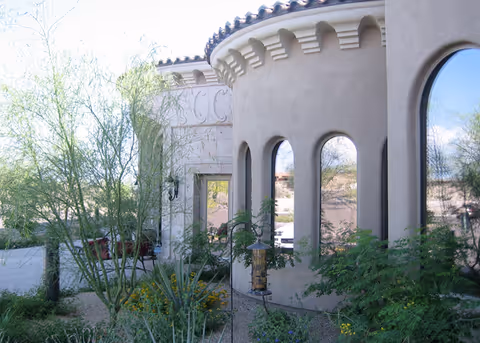 Exterior view of a beige stucco building with arched windows and decorative architectural details, surrounded by desert landscaping including various shrubs and cacti under a clear blue sky.