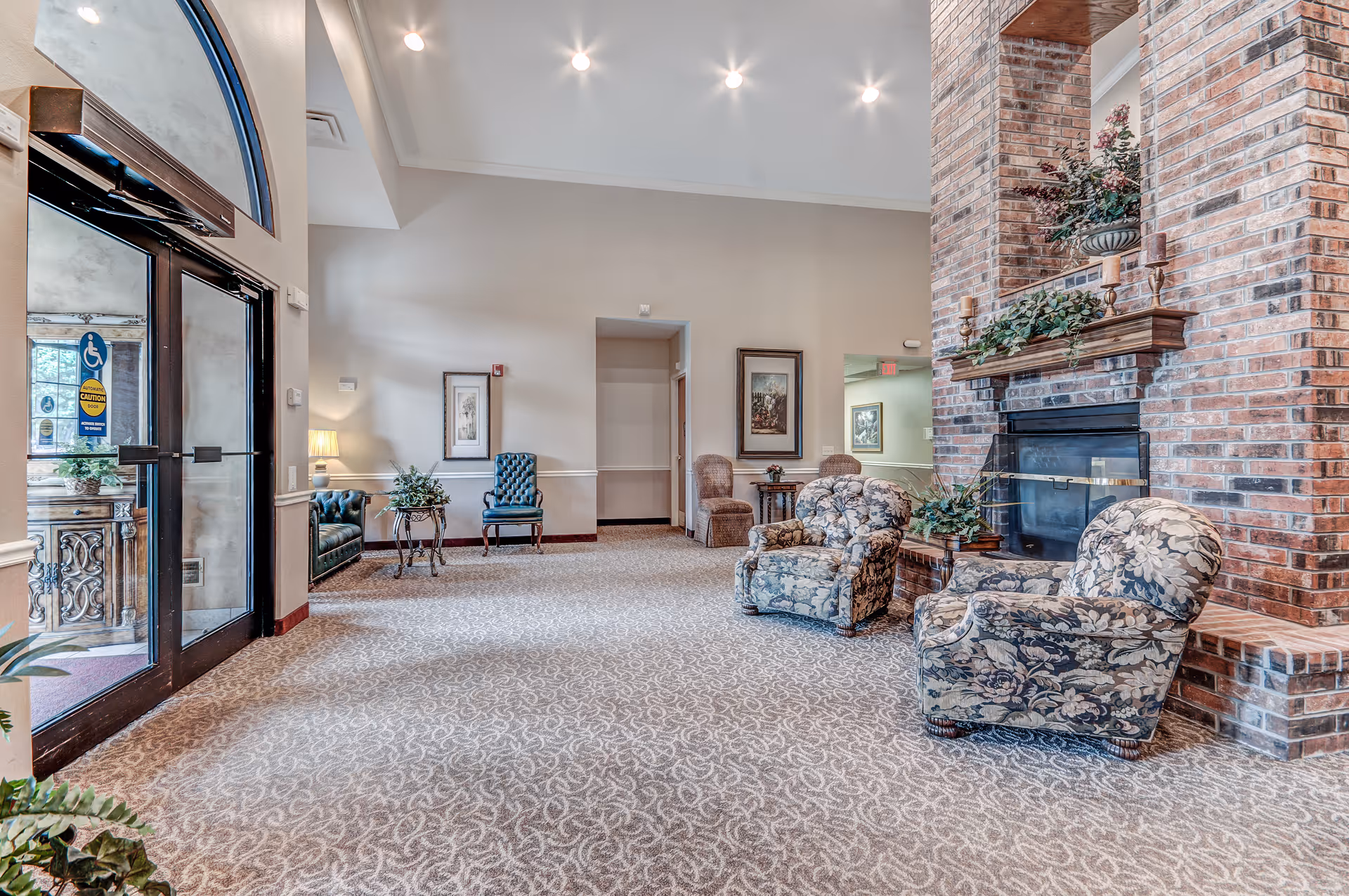 A spacious senior living facility lobby with patterned carpet, floral upholstered armchairs, a large brick fireplace decorated with plants and candles, and a glass entrance door with accessibility signs. The walls are light-colored with framed artwork and additional seating including a blue chair and a small table with a lamp.