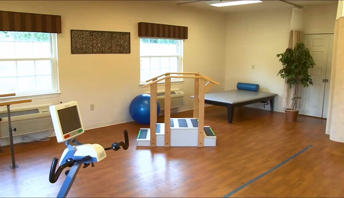 A bright therapy/exercise room with wooden handrails, an exercise bike, a therapy table, and a stability ball on hardwood floors.