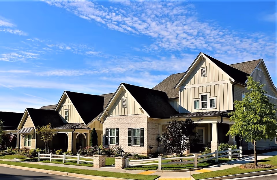 Exterior view of a large, modern residential building with multiple peaked roofs, cream-colored siding, and brick accents. The building is surrounded by a well-maintained lawn, small trees, shrubs, and a white wooden fence. The sky is clear with scattered clouds.