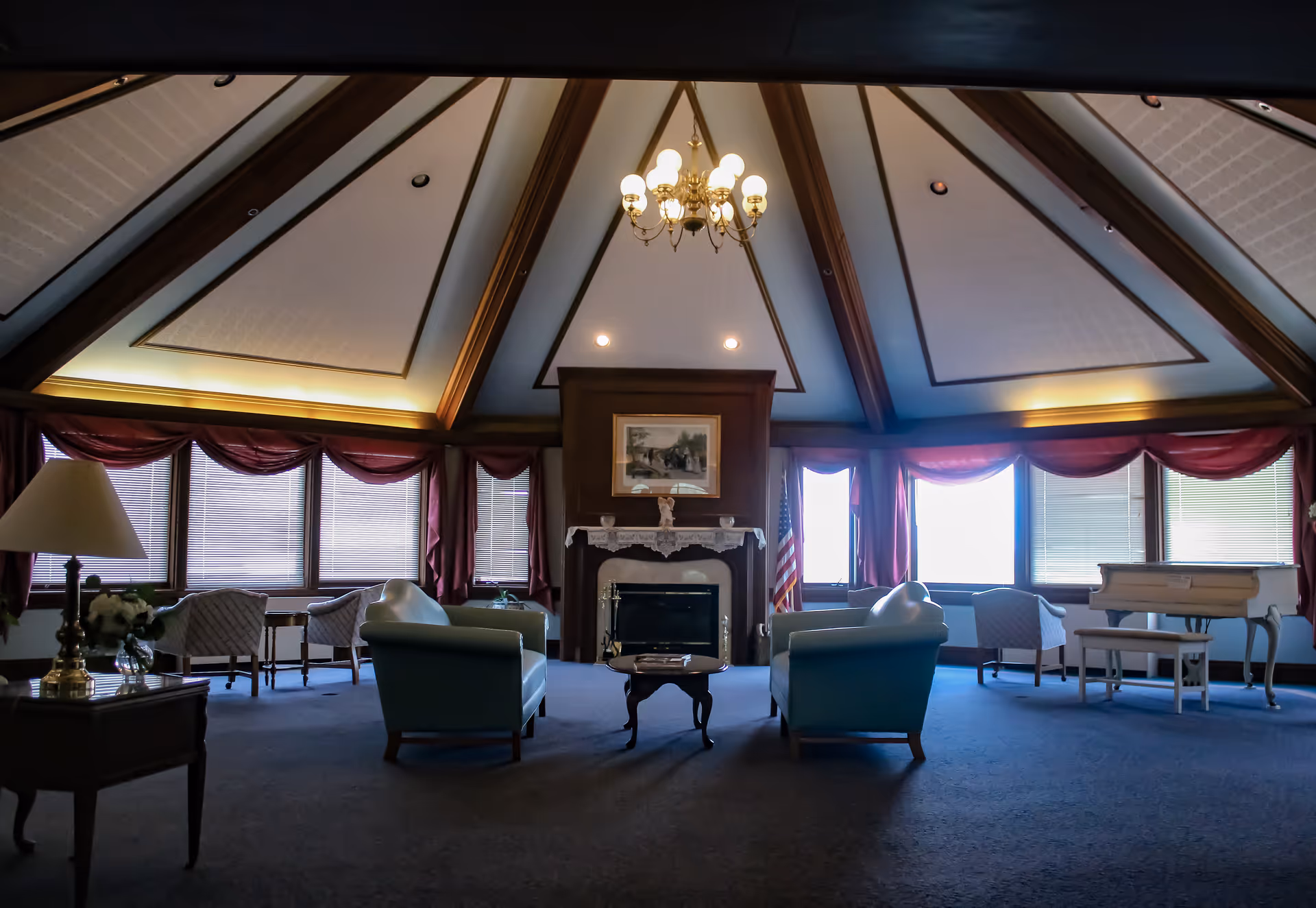 A spacious living room with a high vaulted ceiling featuring wooden beams and a chandelier. The room has large windows with red curtains, several armchairs arranged around a small coffee table, a fireplace with a framed picture above it, a side table with a lamp and flowers, and a white piano with a matching bench.