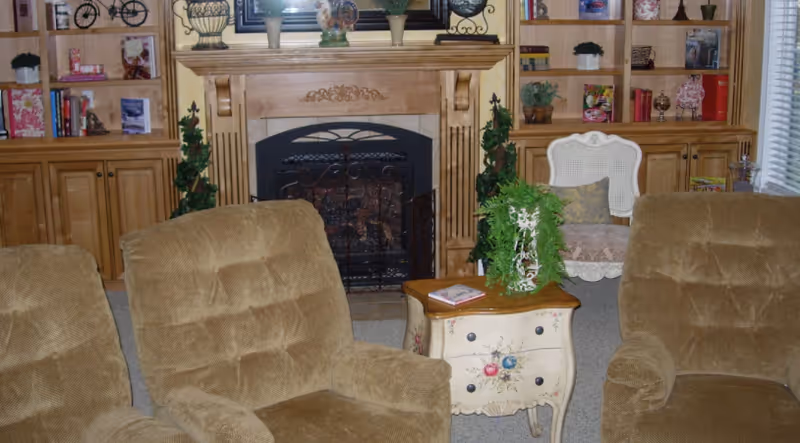 A cozy living room with two brown cushioned armchairs facing a small decorative wooden table with a potted plant and a book on it. Behind the table is a fireplace with an ornate wooden mantel, flanked by built-in wooden shelves filled with books, plants, and decorative items. A white chair with a cushion is placed near the window with blinds.