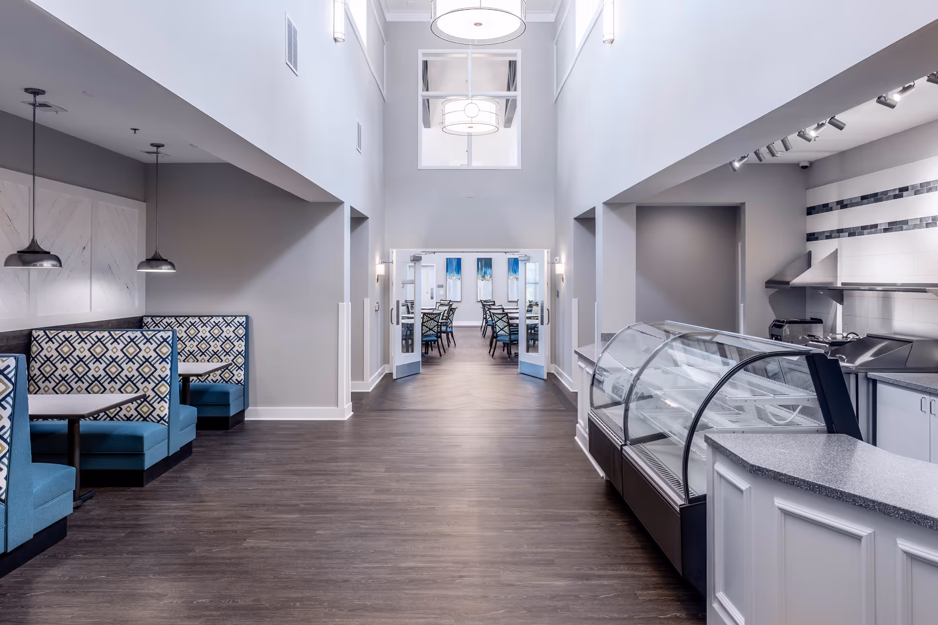 Interior view of a senior living facility dining area with blue patterned booth seating on the left, a glass display case and kitchen area on the right, and an open doorway leading to a room with tables and chairs in the background. The space features high ceilings, modern light fixtures, and wood flooring.