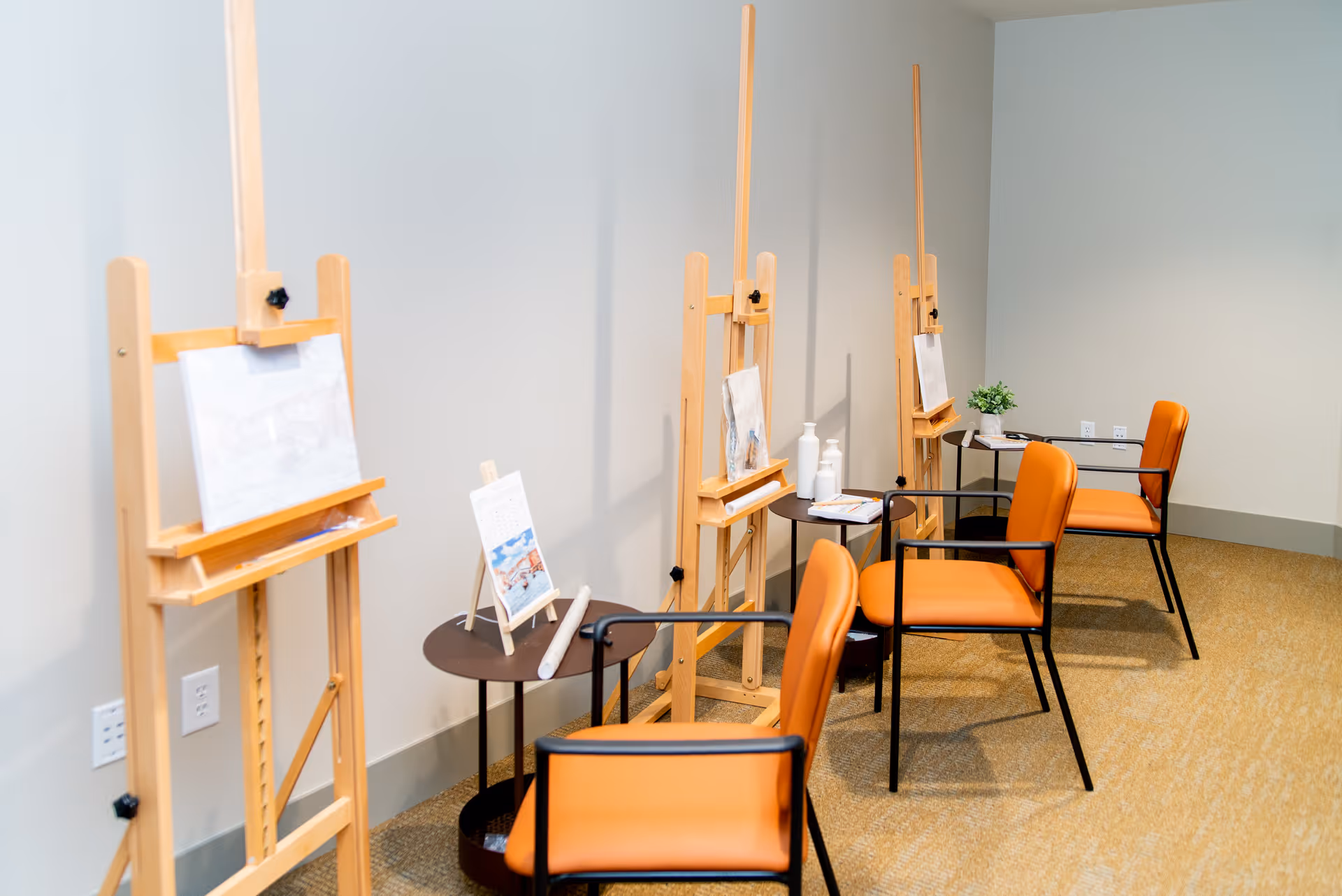 A room with three wooden easels holding canvases, each paired with an orange chair and a small round table. The tables have art supplies and a small plant, suggesting an art or painting activity area.