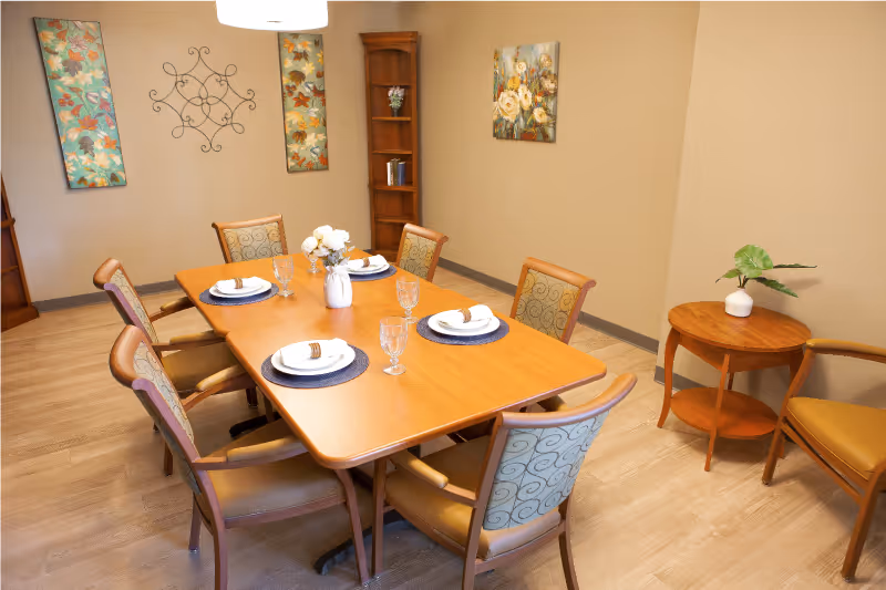 A dining room with a rectangular wooden table set for six people with plates, napkins, and glasses. The room has six chairs with patterned upholstery around the table. There are three floral paintings and a decorative metal wall art on the beige walls. A small round wooden side table with a potted plant is placed near the wall.