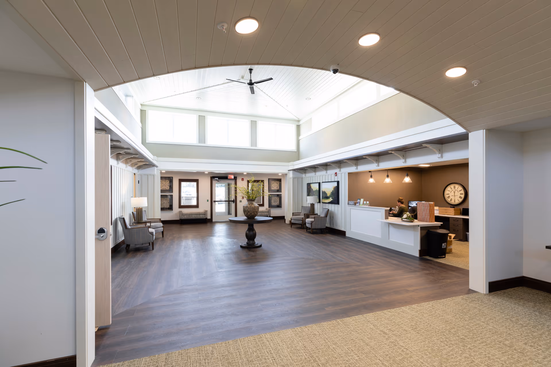 Spacious and well-lit senior living facility lobby with high ceilings, large windows, and a ceiling fan. The room features a central round table with a decorative vase, seating areas with chairs and lamps along the walls, and a reception desk with a staff member working behind it. The flooring transitions from carpet to wood, and the overall decor is modern and welcoming.