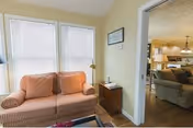 A cozy living area with a peach-colored loveseat featuring patterned cushions, positioned near two large windows with closed blinds. Next to the loveseat is a small wooden side table with a lamp and a framed picture on the wall above it. Through an open doorway, another room is visible with additional seating including a sofa and a lamp, suggesting a comfortable common living space.