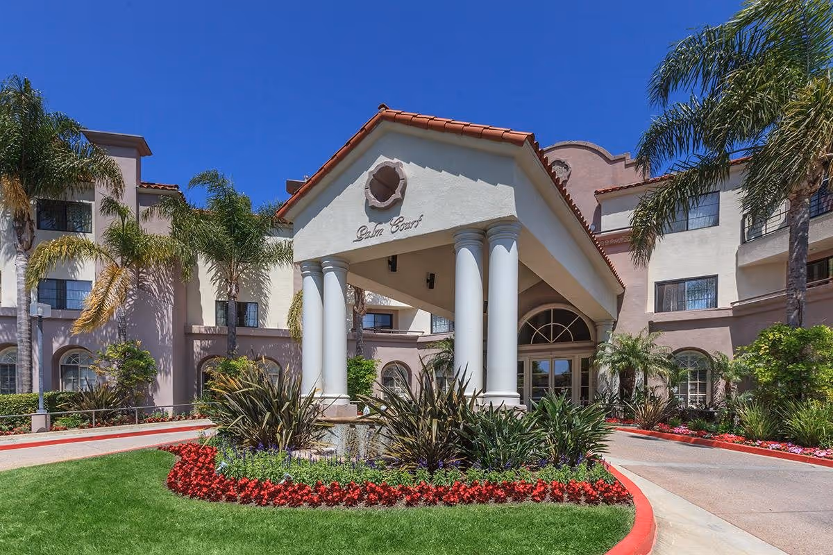 Exterior view of Palm Court facility entrance with a covered driveway supported by white columns, surrounded by palm trees, green grass, and colorful flower beds under a clear blue sky.