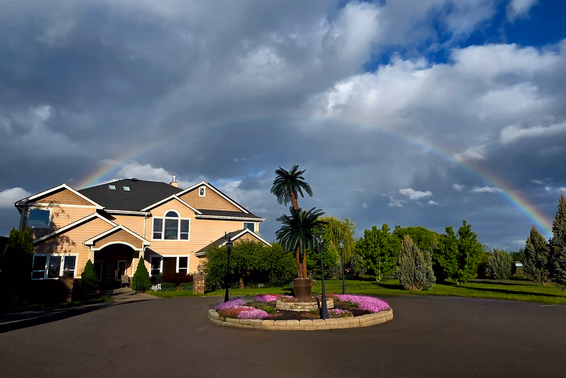 A large two-story house with beige siding and a dark roof under a cloudy sky with a visible rainbow arching over it. In front of the house is a circular driveway with a landscaped island featuring palm trees and pink flowers. Surrounding the area are green trees and shrubs.