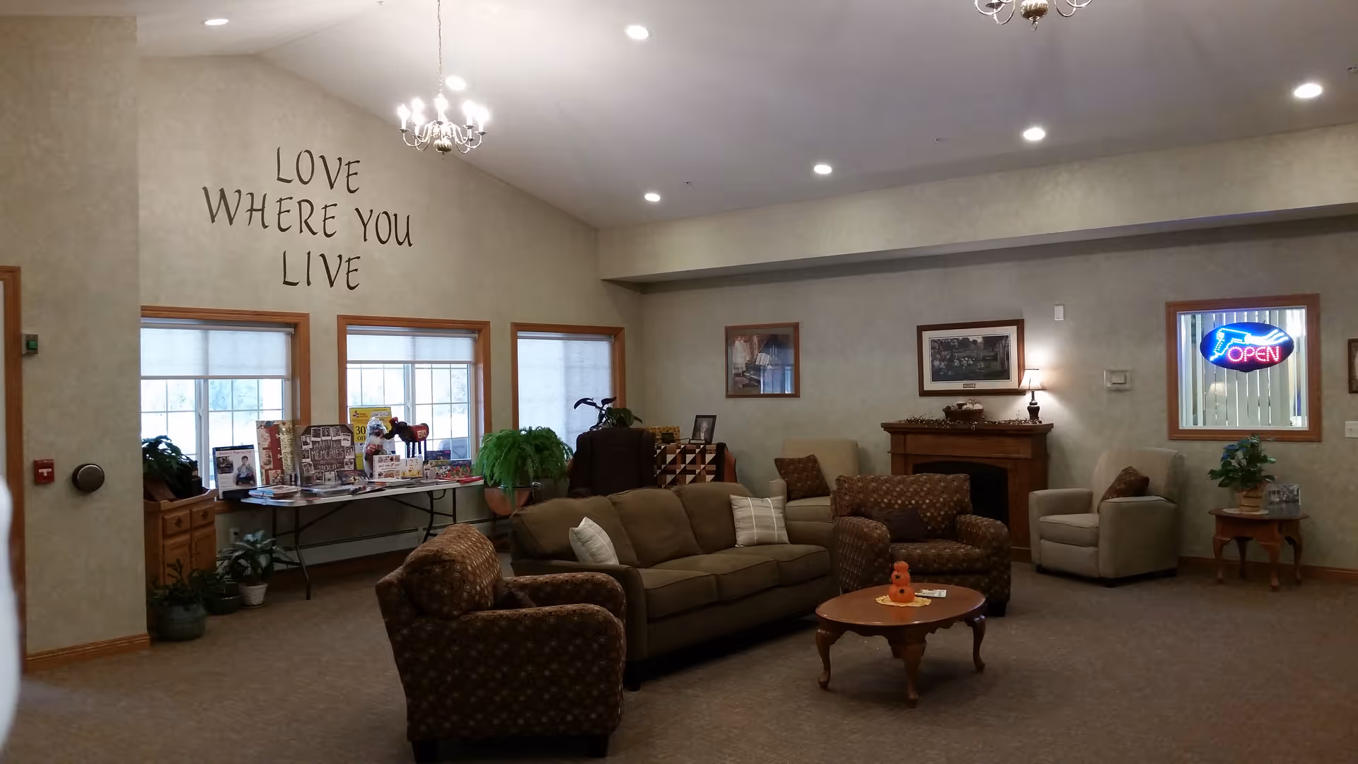 A cozy living room area in a senior living facility with a sofa, armchairs, a coffee table, and a fireplace. The walls are decorated with framed pictures and a large wall decal that reads 'LOVE WHERE YOU LIVE'. There are three windows with blinds, a table with brochures and plants, and a neon 'OPEN' sign in one window.