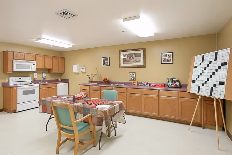 A well-lit room with a kitchen area featuring a stove, microwave, dishwasher, and wooden cabinets. In the center, there is a table covered with a checkered tablecloth and two chairs. On the right side, there is a large crossword puzzle board on an easel. The walls are decorated with framed pictures.