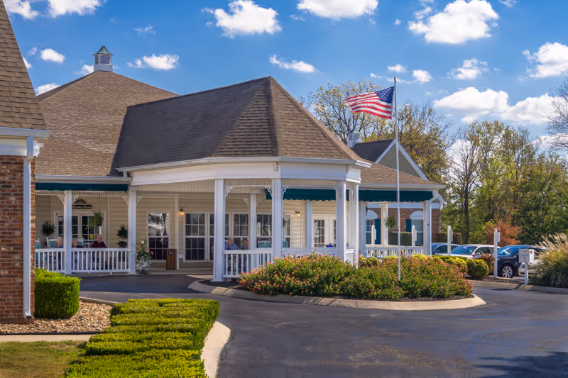 Front entrance of a senior living facility with a covered porte-cochere, American flag, and landscaped circular driveway.